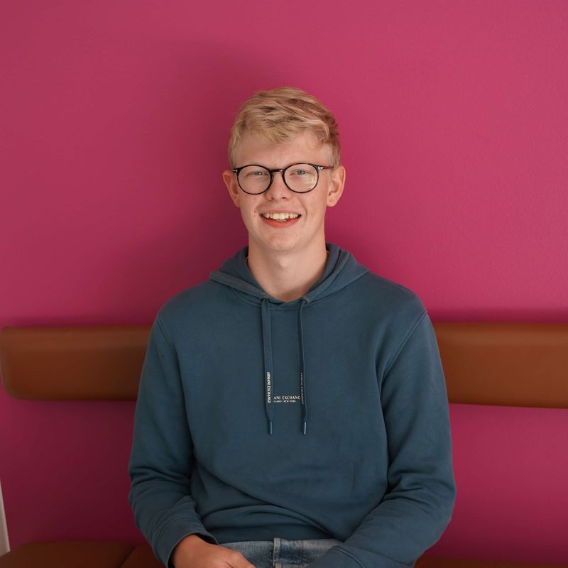 Isak ler mot kameran, sitter på bänk framför rosa vägg