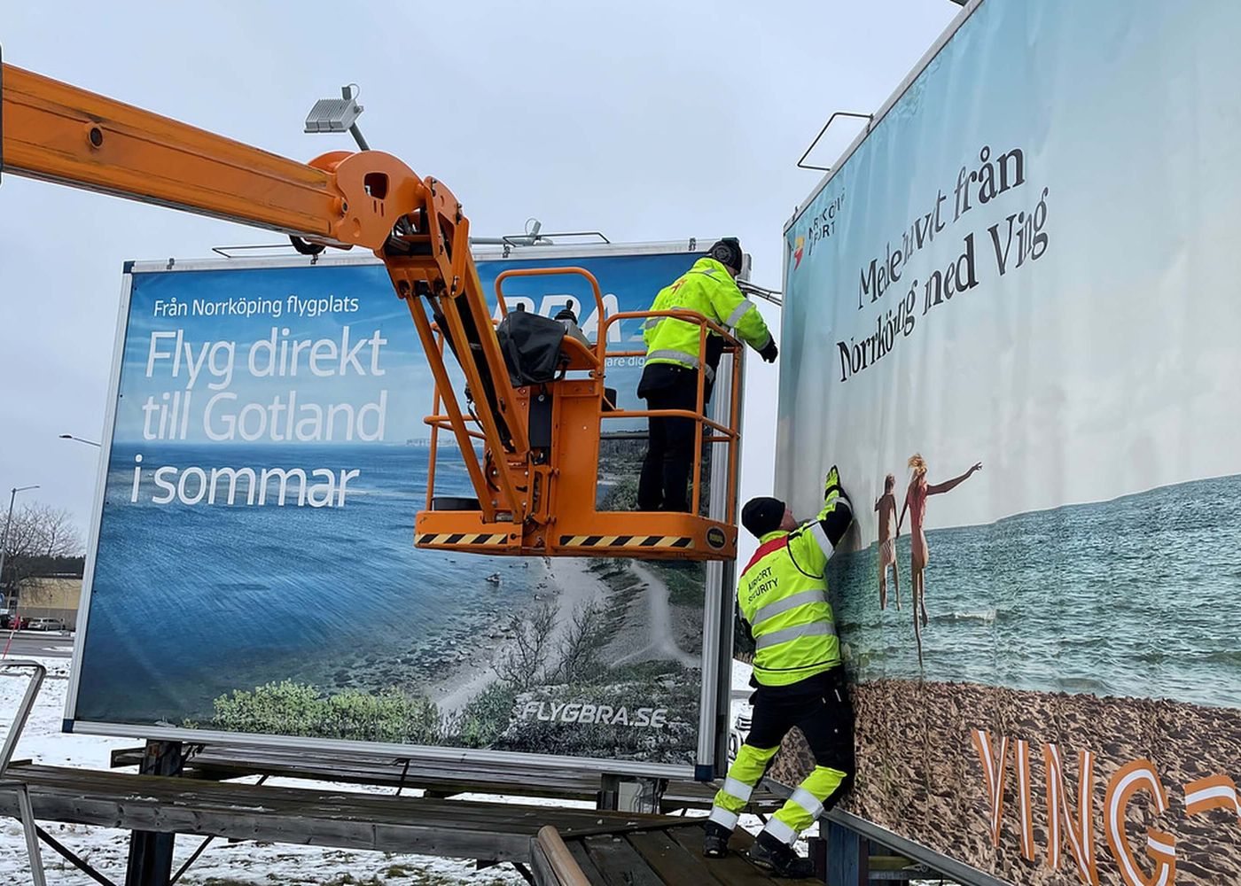 Två personer i varselkläder sätter upp reklamaffischer vid Norrköpings flygplats, en med texten 'Flyg direkt till Gotland i sommar'