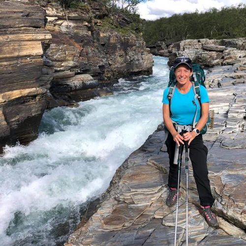 Kvinna med ryggsäck står leende på klippor vid en forsande bergsälv under vandring.