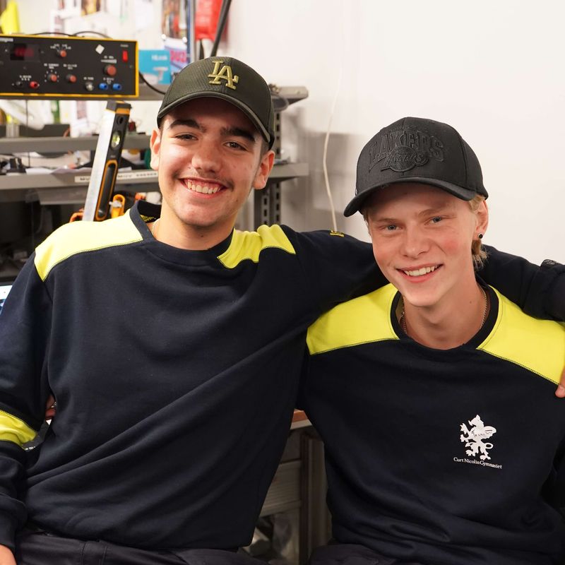 Wiggo och Emir håller om varandra och ler mot kameran i elsalen på CNG