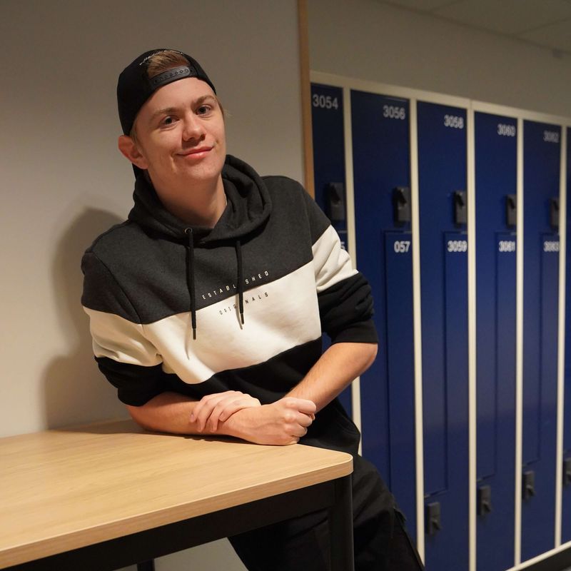 Alexander ler mot kameran, lutad mot ett bord i korridoren på CNG