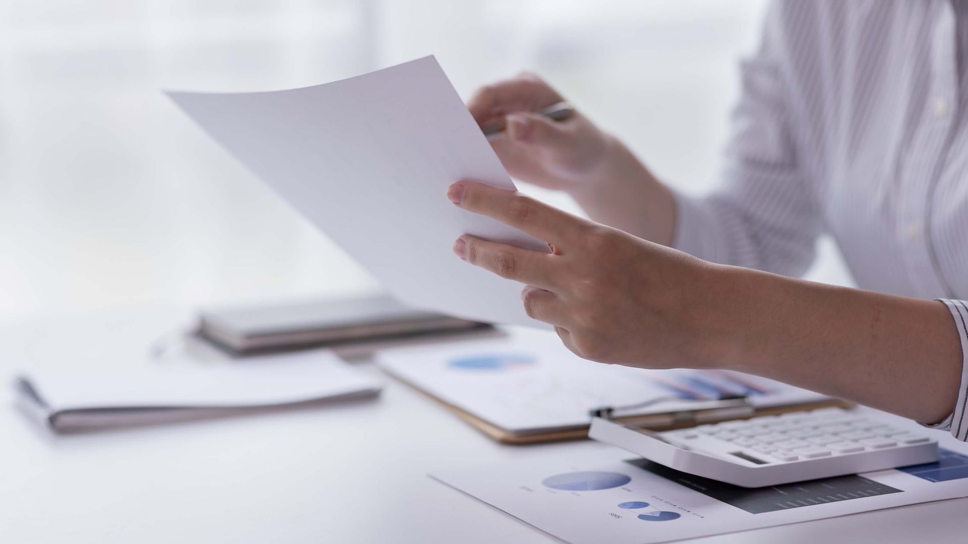 Närbild på händer med papper och penna. På bordet ligger ekonomipapper med grafer och en miniräknare.