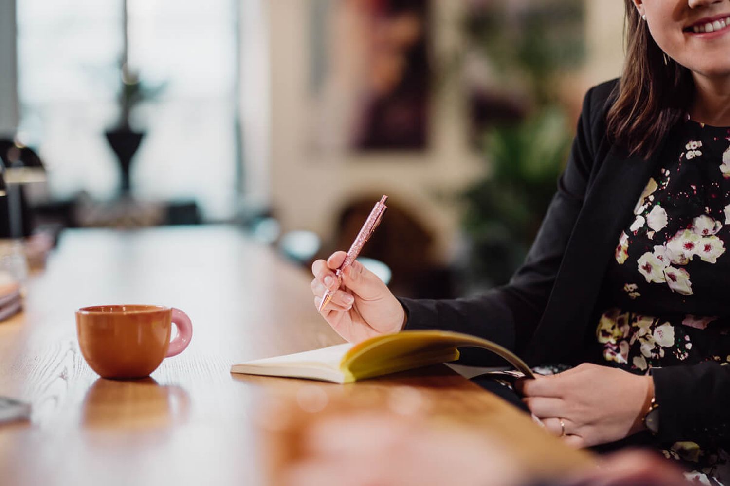 Person writing in a notebook with a coffee cup on the table, blurred background
