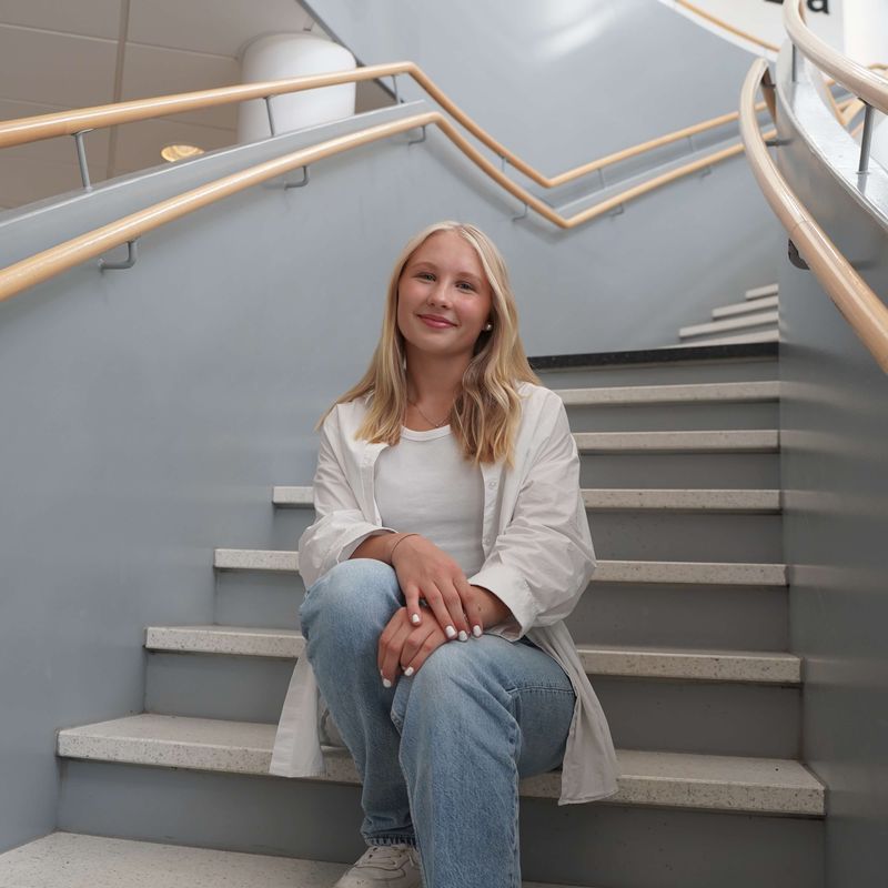 Engla ler mot kameran, sitter i trappan på skolan
